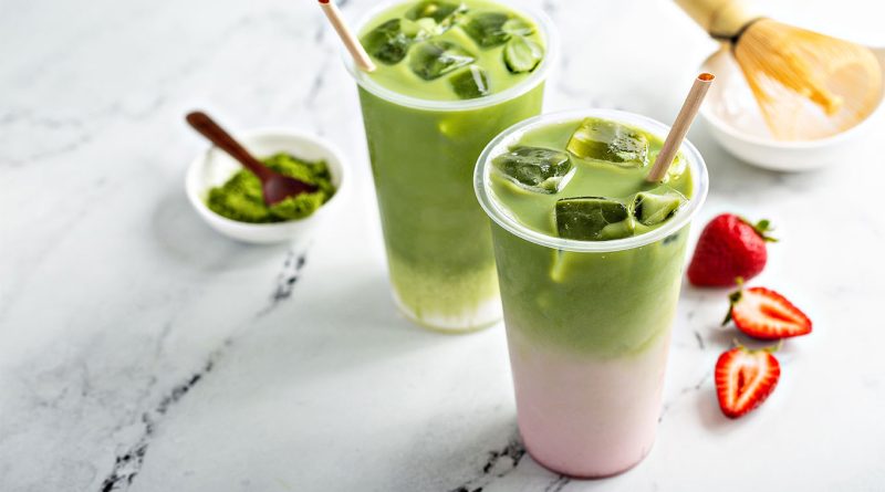 Two iced matcha drinks with pink and white milk layers served in clear cups, placed on a marble surface with fresh strawberries, a bowl of matcha powder, and a bamboo whisk in the background