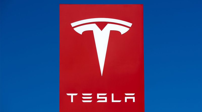 Tesla logo on a bright red sign against a clear blue sky, representing the brand’s presence and identity
