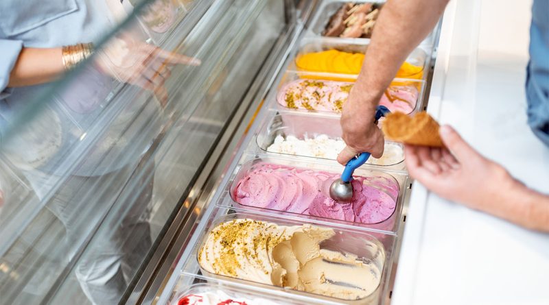 A server scoops naturally colored ice cream into a cone at a display counter, showcasing a variety of pastel-toned flavors without artificial dyes.