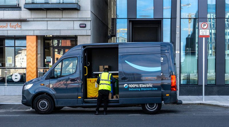 Amazon delivery worker loads packages into a 100% electric delivery van parked in an urban area outside a commercial building.