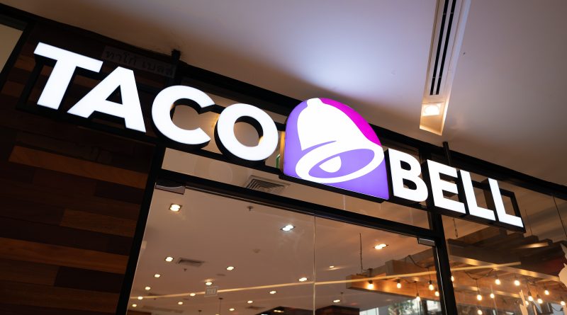 Illuminated Taco Bell restaurant sign above a glass storefront at night, with warm interior lighting visible inside