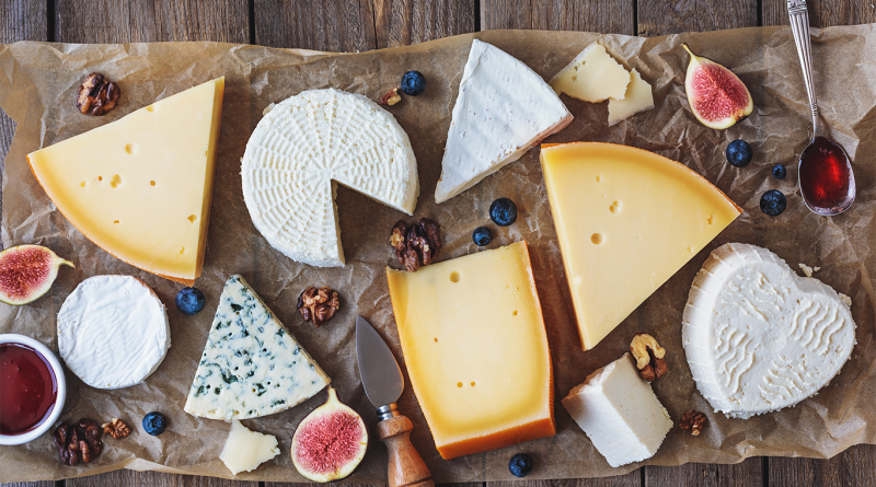A rustic cheese board featuring a variety of leftover cheeses including brie, blue cheese, ricotta, and hard cheeses, surrounded by figs, blueberries, walnuts, and a small knife on parchment paper.