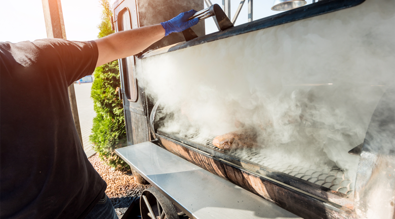 A person opens a smoking barbecue grill outdoors with thick smoke billowing out, representing traditional barbecue cooking