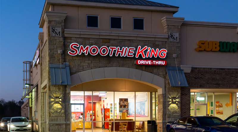 Exterior of a Smoothie King storefront at dusk, with illuminated “Smoothie King” and “Drive-Thru” signage and a Subway sign visible next door.