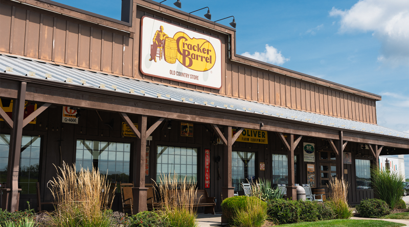 Exterior view of a Cracker Barrel Old Country Store with a large wooden facade, front porch with rocking chairs, and landscaped greenery under a blue sky.