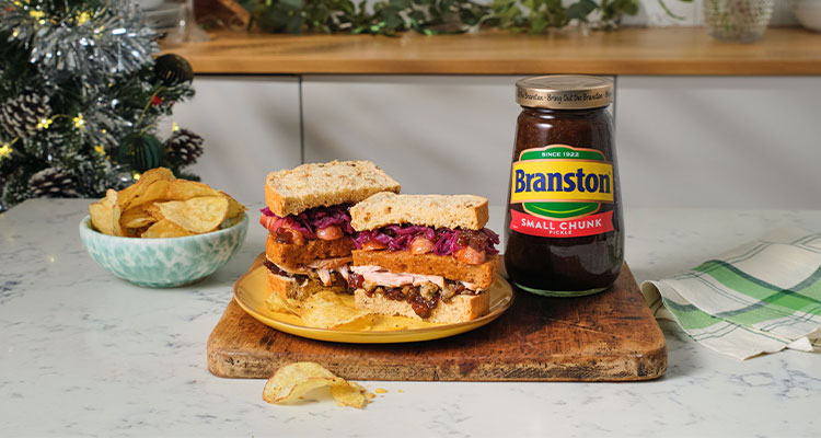 a jar of Branston Small Chunk Pickle alongside two hearty sandwiches and a bowl of crisps