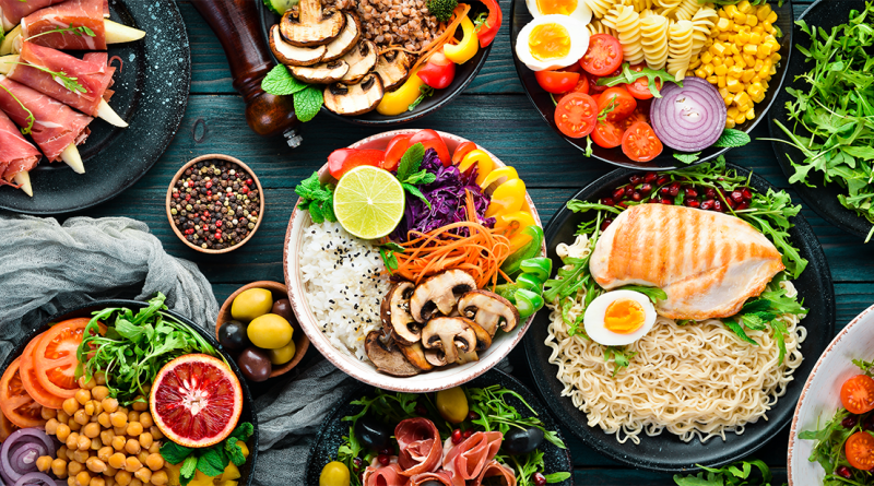 Overhead view of healthy meal bowls with grilled chicken, rice, mushrooms, pasta, eggs, chickpeas, fresh vegetables and leafy greens arranged on a wooden table