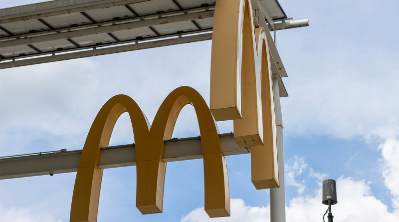 McDonald’s golden arches sign mounted beneath solar panels against a blue sky with clouds, photographed from below.